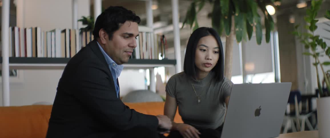 A man and a woman are sitting on a sofa and are talking while they are wearing formal clothes and in front of them there is a laptop