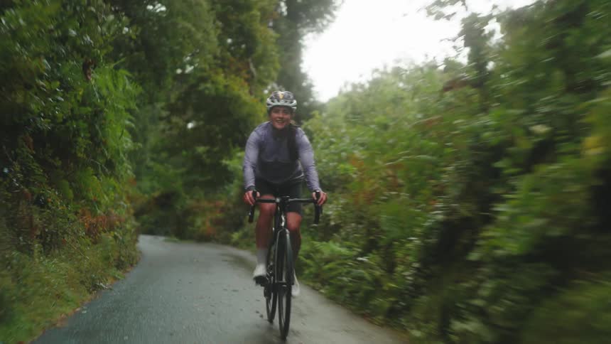 Female cyclist smiles while pedaling along paved path through the woods