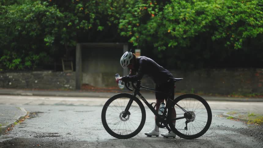 A cyclist wearing a helmet is standing on his bike with a bottle of water in his hand and is looking down with a tired look