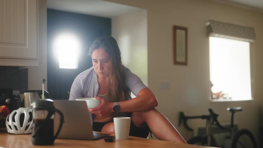 A woman cyclist is eating cereal while looking at her laptop