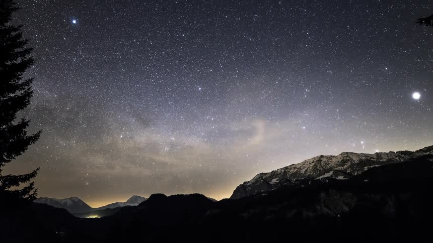 Conifer trees standing near mountains are silhouetted against the night sky