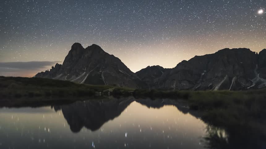 Darkening daytime landscape with imposing mountains under a starry sky reflected in a lake