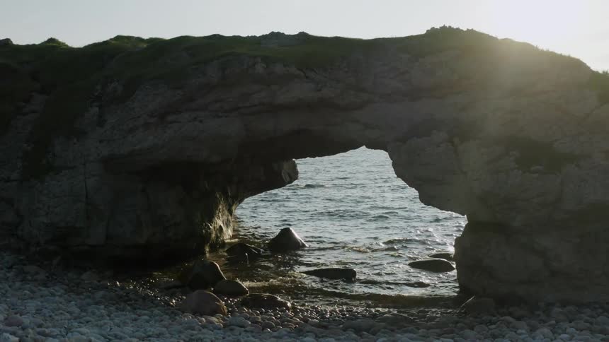 Small waves flow through stone arch to splash against a pebble strewn shore during the day