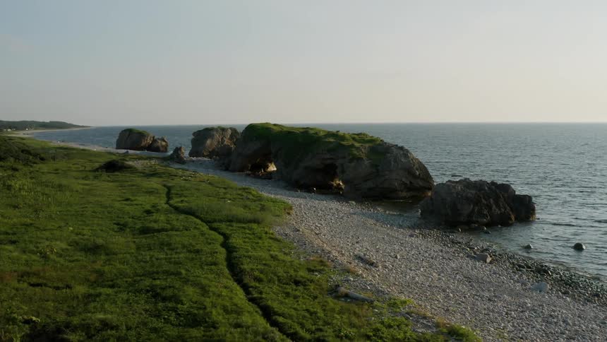 Coastal landscape with a pebble beach large rocks in the sea and a green field under a clear sky