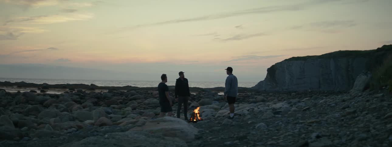 Three people are standing around a fire on a floor with many stones