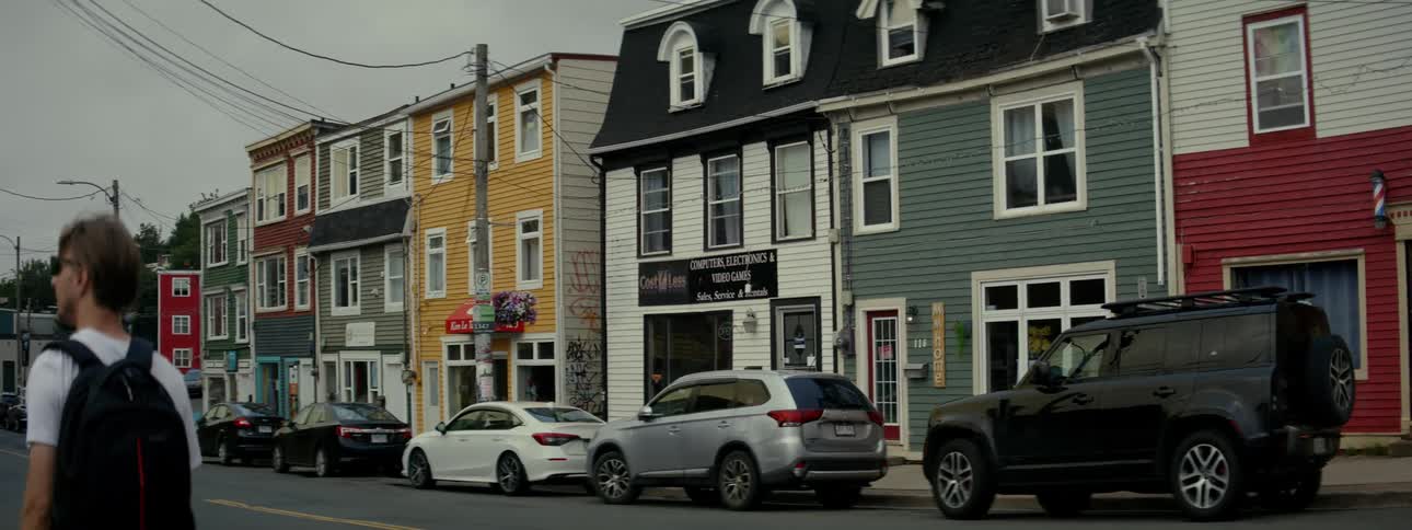 A facade of several colorful twostory buildings with shops at the bottom and several cars parked on the street and a man walking in front