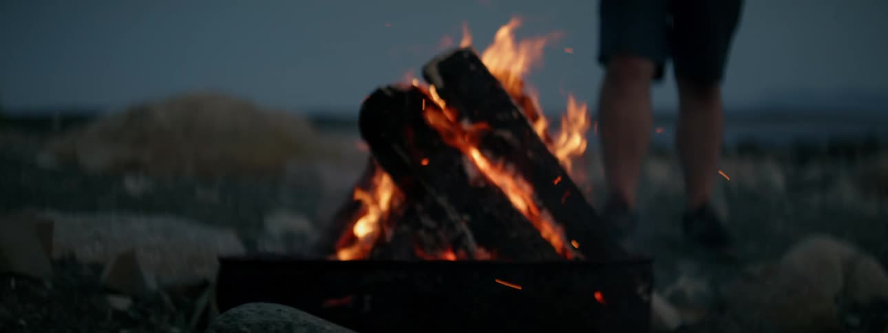 Bonfire lit amidst rocks and a persons legs in the background