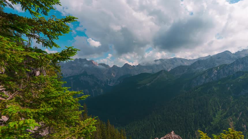 A landscape of a mountain range with the base covered in forest trees and white clouds passing in the sky