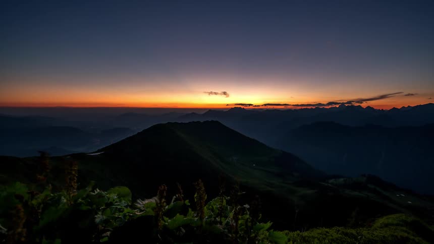 Sunrise seen from the top of a mountain with silhouettes of other mountains in the background and the sky gradient from blue to orange