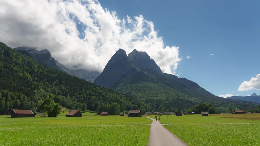 Pedestrians walk along path passing through small town near forested mountains on a cloudy day