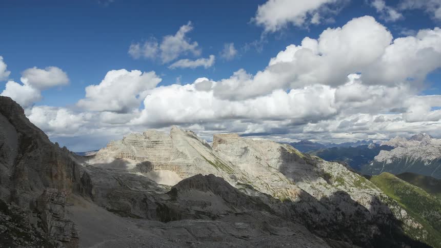 A mountain range with mountains illuminated by sunlight and several white clouds passing quickly in the clear blue sky