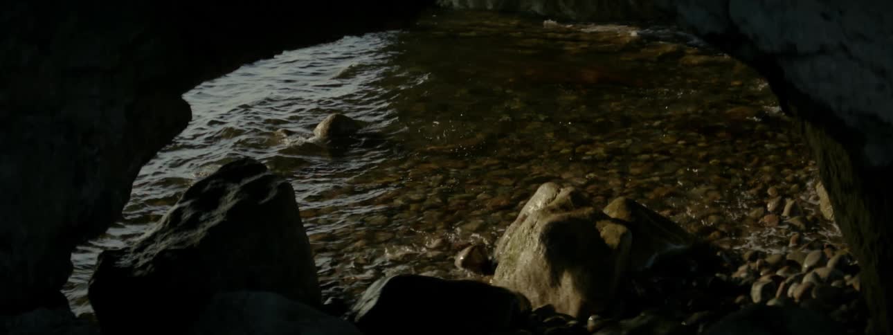 Water laps at boulder and pebble strewn shore near small arching rock formation