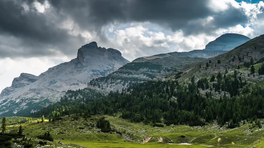 A landscape of a mountain range with trees at the base of the mountains and gray clouds passing in the blue sky