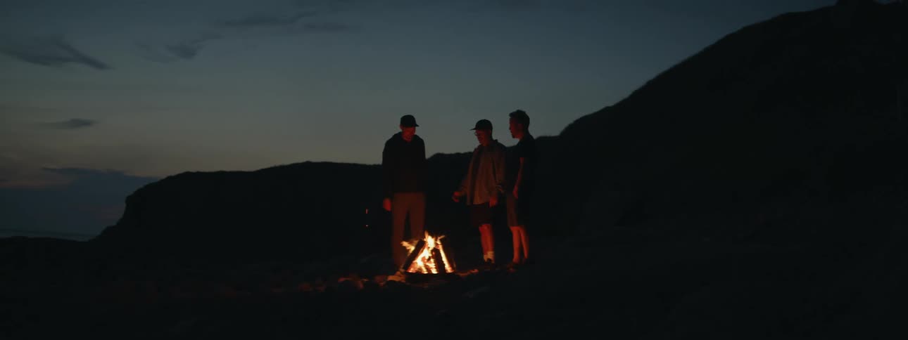 Three men stand next to a campfire in the mountains at twilight