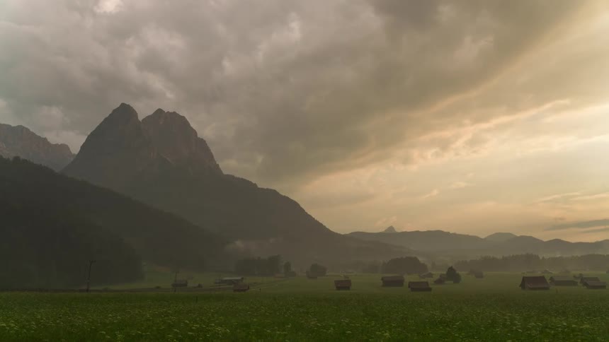 Vast green field with scattered structures under a dramatic sky with mountains in the background