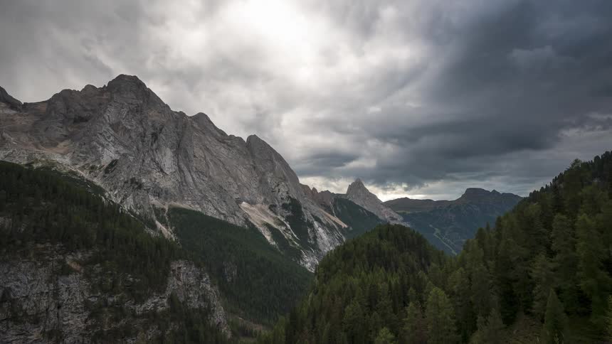 A landscape of a mountain range and on the left side there is a pine forest and in the sky white clouds passing quickly