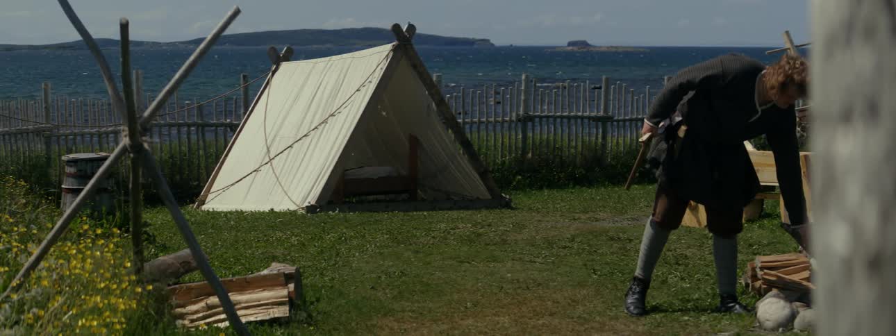 A man putting a piece of firewood on a fire and another man watching and both are in a place with a tent at a campsite
