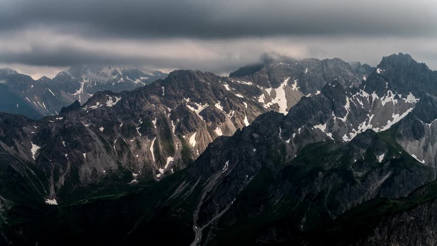 A mountain range with snow on the surface of some mountains and clouds passing quickly in the sky