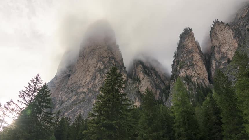 Sun peeks around a cluster of narrow mountains towering over a conifer forest on a cloudy day