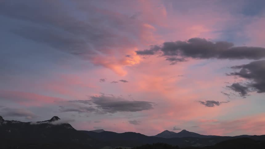 A landscape of a mountain range and the sky is orange and dusk while some clouds pass by