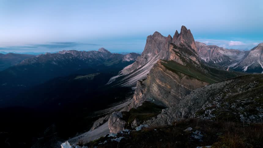Clouds flow and wind blows grass as the sun lights up the slope of a mountain before going dark