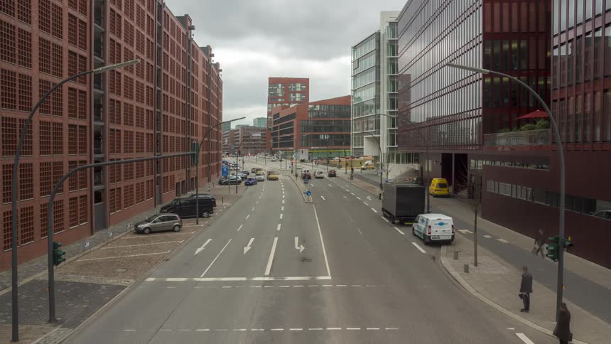 Modern urban scene with multiple lanes of traffic and moving vehicles flanked by tall red and brown buildings under a cloudy sky