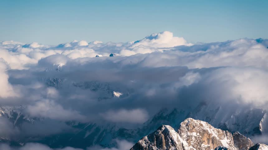 Several dense white clouds passing above snowcovered mountains in a mountain range