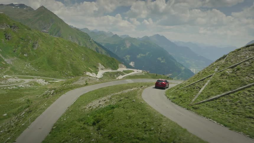 Red vehicle travels along road winding across green mountains overlooking a foggy valley
