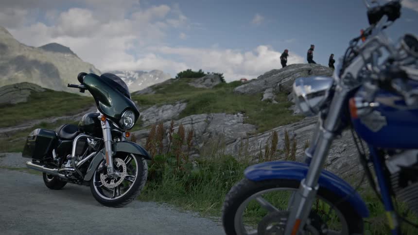 Two parked motorcycles in the foreground with three people walking in the background against a mountainous backdrop