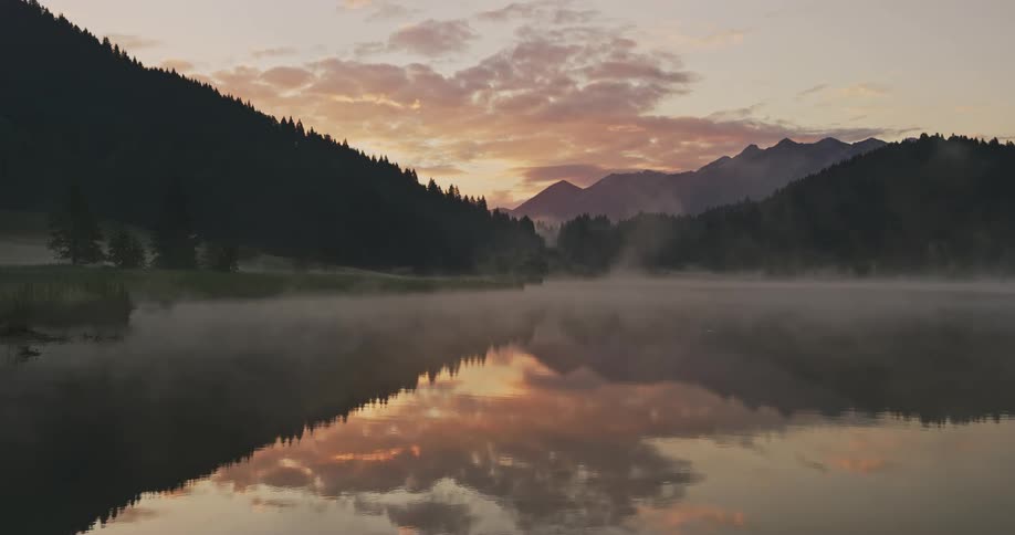 Lake waters reflect sun rising over forested mountains
