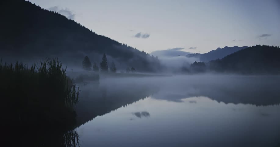 Fog flows across lake water reflecting forested mountains on a cloudy day