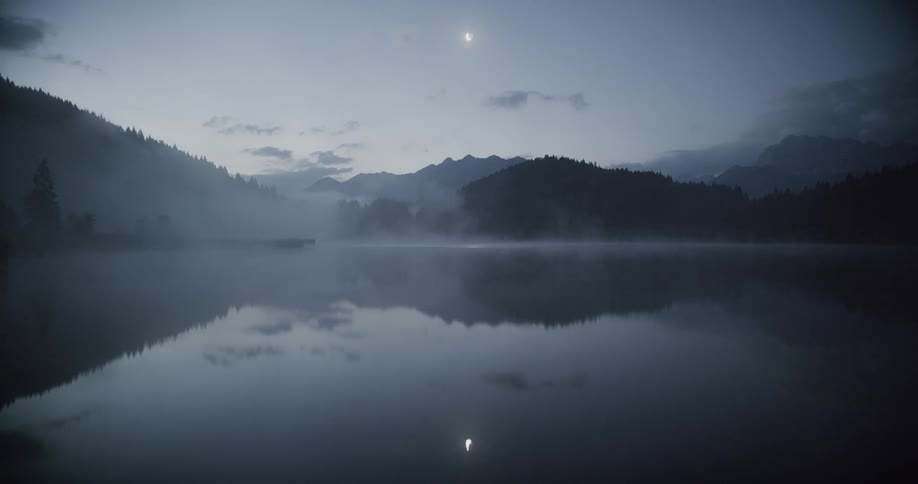 A lake reflecting mountains of trees around it and the moon in the sky shining brightly