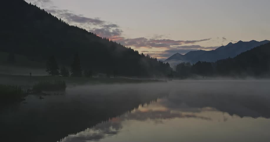 A lake with gentle fog overhead in a region with treecovered mountains
