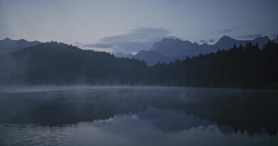 Landscape of a lake with mist reflecting mountains and conifers around