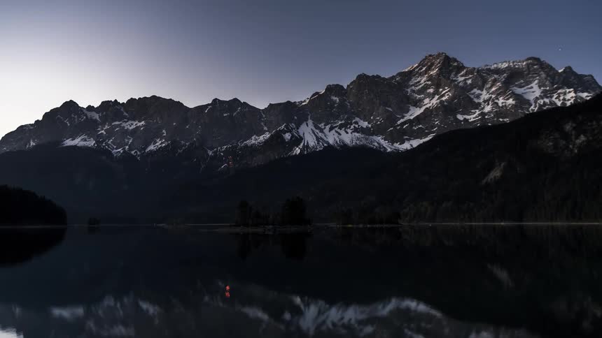 Rising sun tints sky obscuring stars over reflective lake and nearby snow dusted mountains