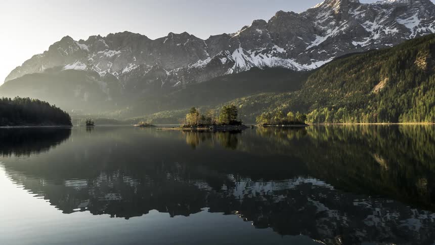 Landscape of a lake reflecting the surrounding snowy mountains and conifers on a morning