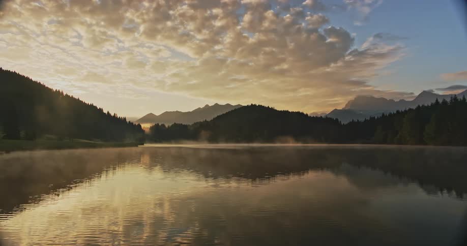A misty lake with a reflection of the sky and clouds flanked by silhouetted mountain ranges during sunrise or sunset