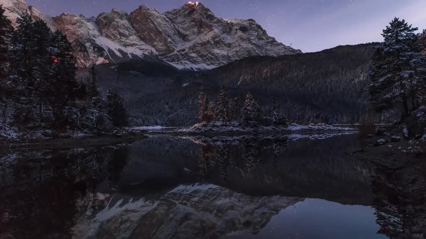Cars driving near a lake and mountains light up the surrounding area as night falls