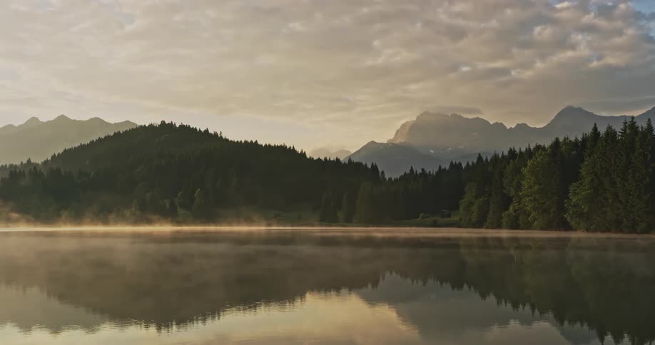 A lake with a small mist on its surface and the water on the side is reflecting two secret hills of pine trees