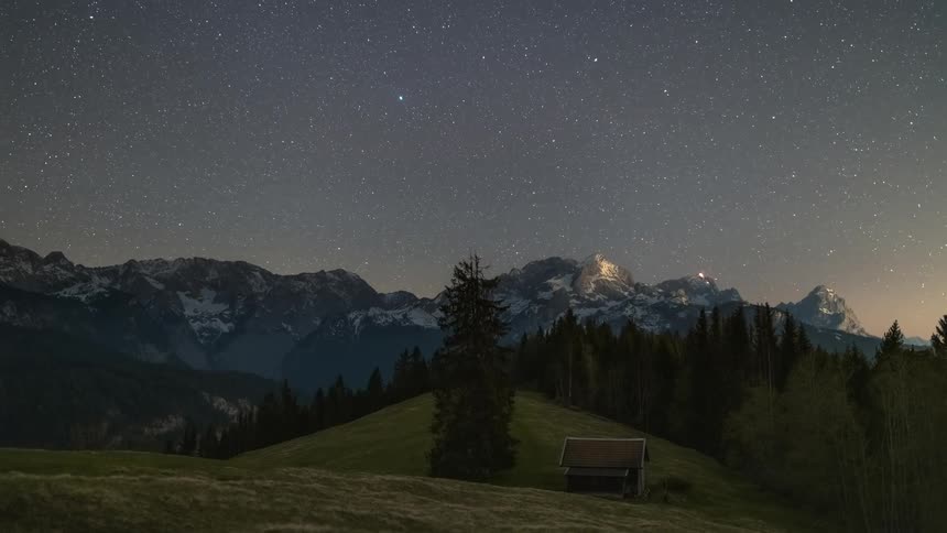 Landscape of a field with conifers and snowy mountains in the background against a sky with stars and the dawning day