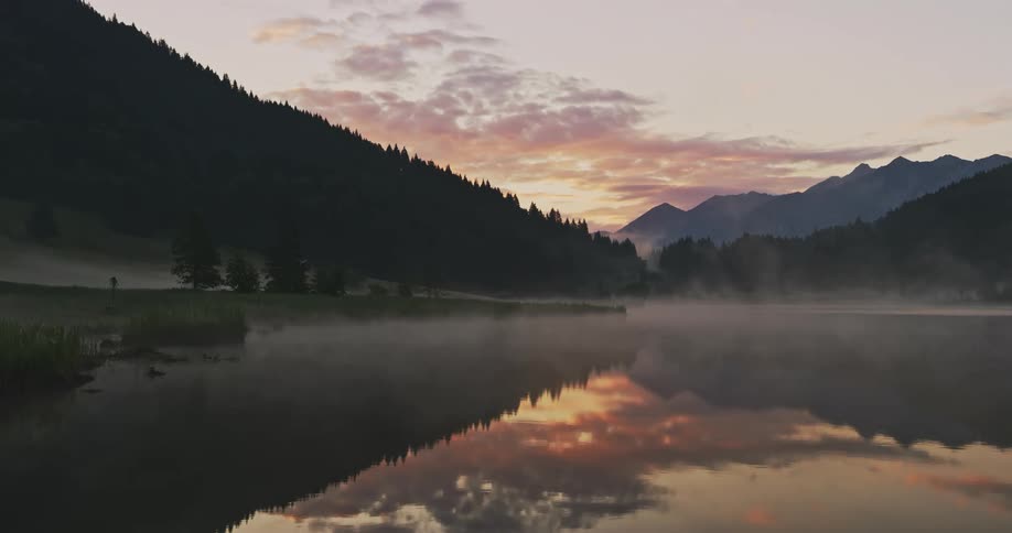 A lake with fog passing over its surface and the water is reflecting the landscape of mountains with trees