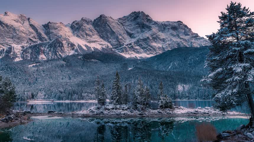 Natural landscape of a lake surrounded by mountain ranges and snowy trees against a purple sky