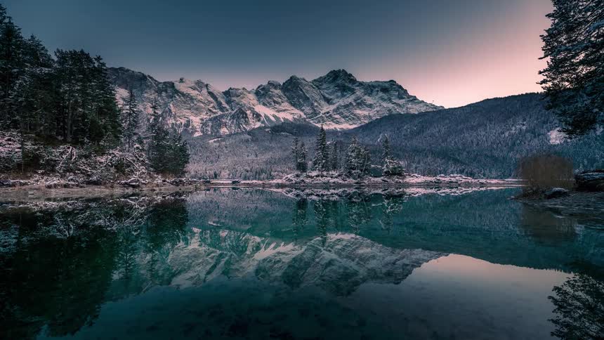 A lake with crystal clear water reflecting the sky and a mountain that is covered in snow and is getting dark