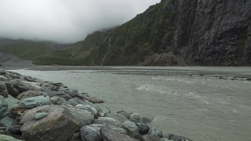 River waters flow between a mountain cliff and a boulder strewn slope on an overcast day