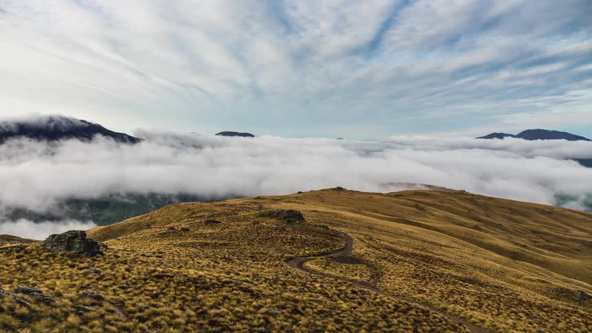 Natural landscape with a winding path that runs through a field of yellowed grass under a dramatic sky with low clouds and mountains in the background