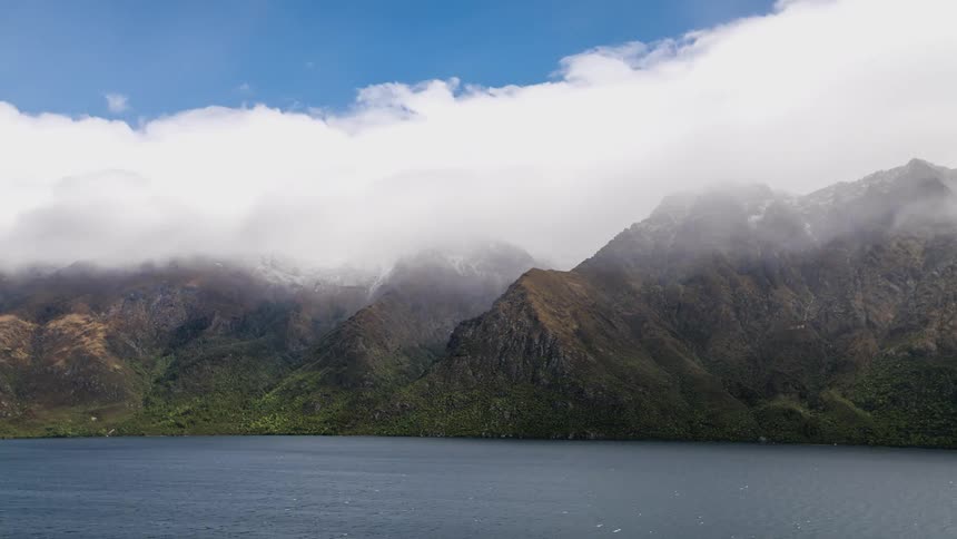 Several dense white clouds passing over the peak of a mountain range near the lake