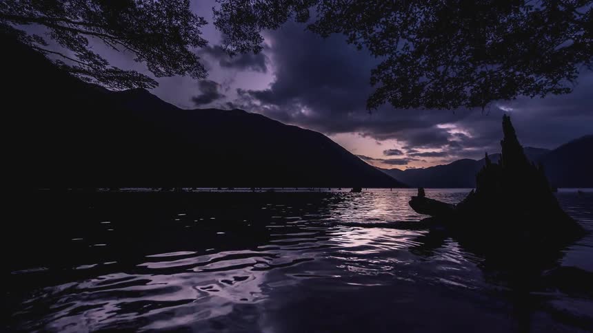 Water flows in a lake while clouds float over nearby mountains