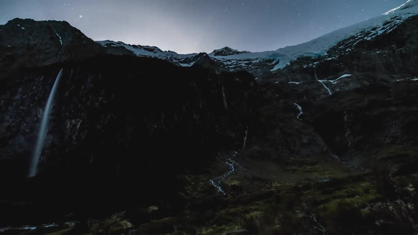 Starry night sky replaces the sun as day turns to night for snowy mountains containing a waterfall