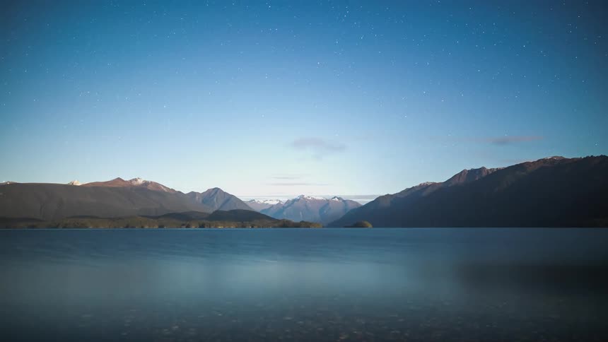 Watercraft crosses water passing mountains silhouetted under a starry sky before night turns into day