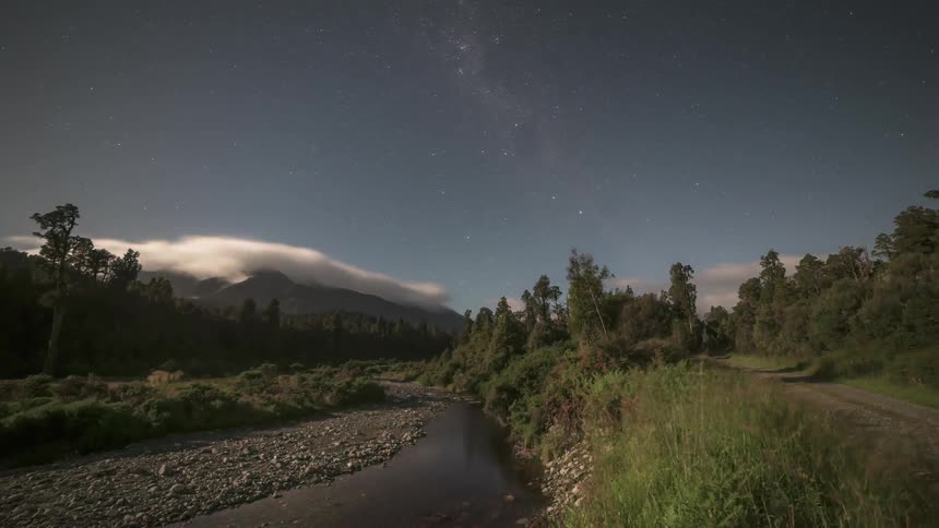 Night landscape with a starry sky above a riverbed surrounded by vegetation and mountains in the background with the day dawning